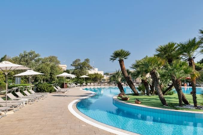 VOI Floriana Resort baseina līkumi un sauļošanās terase ar baltiem saulessargiem — palmas un zaļš dārzs fonā
