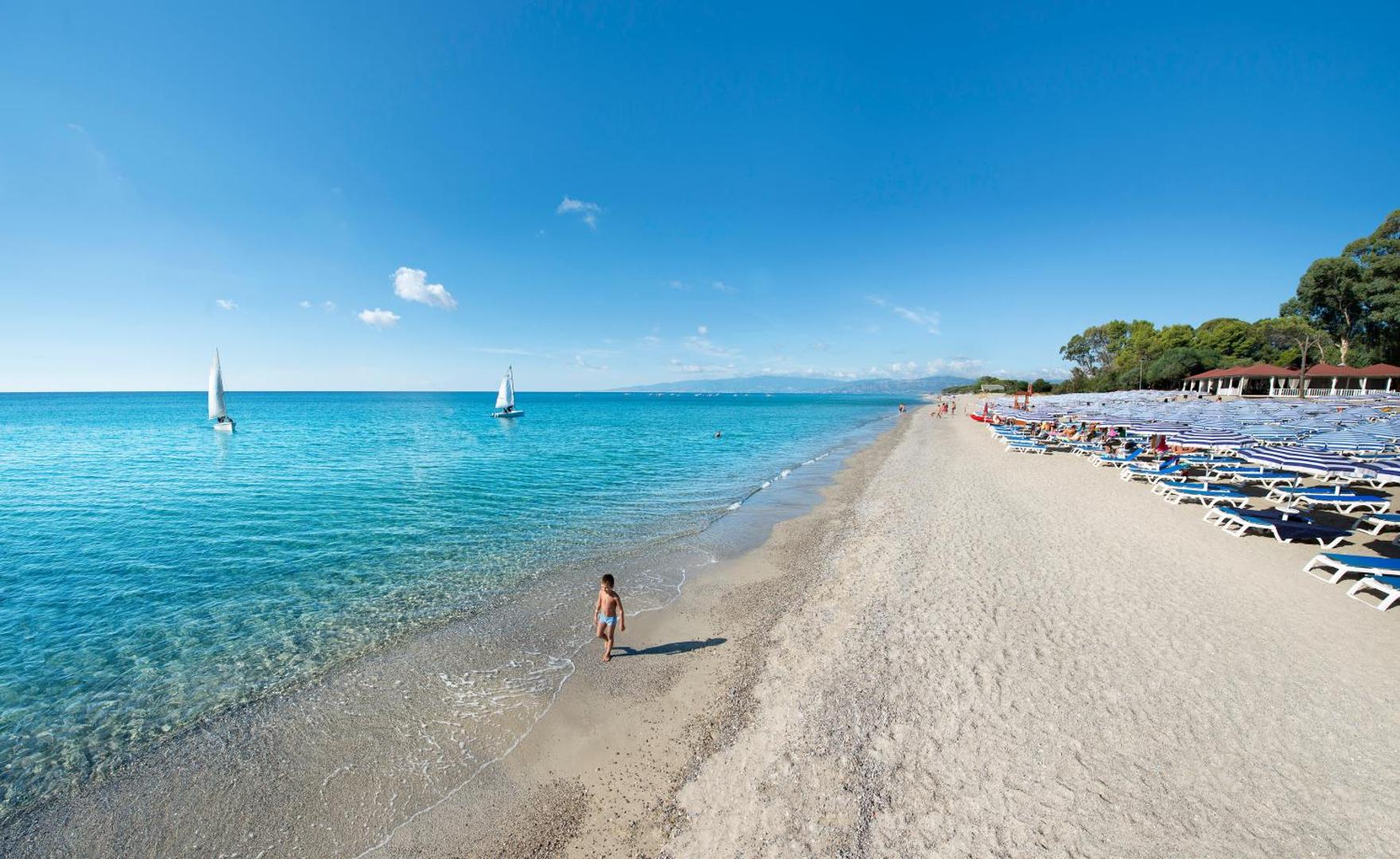 VOI Floriana Resort plašā pludmale ar zili svītrainiem saulessargiem, bērnu un buru laivām tālumā Kalabrijas piekrastē