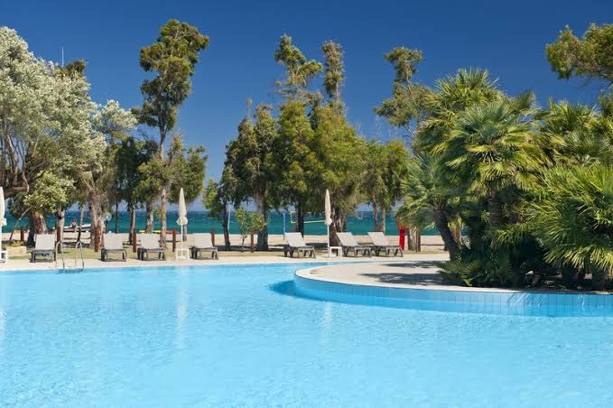 Skats no VOI Floriana Resort baseina uz Jonijas jūras piekrasti — pludmales krēsli zem palmām un zilas debesis