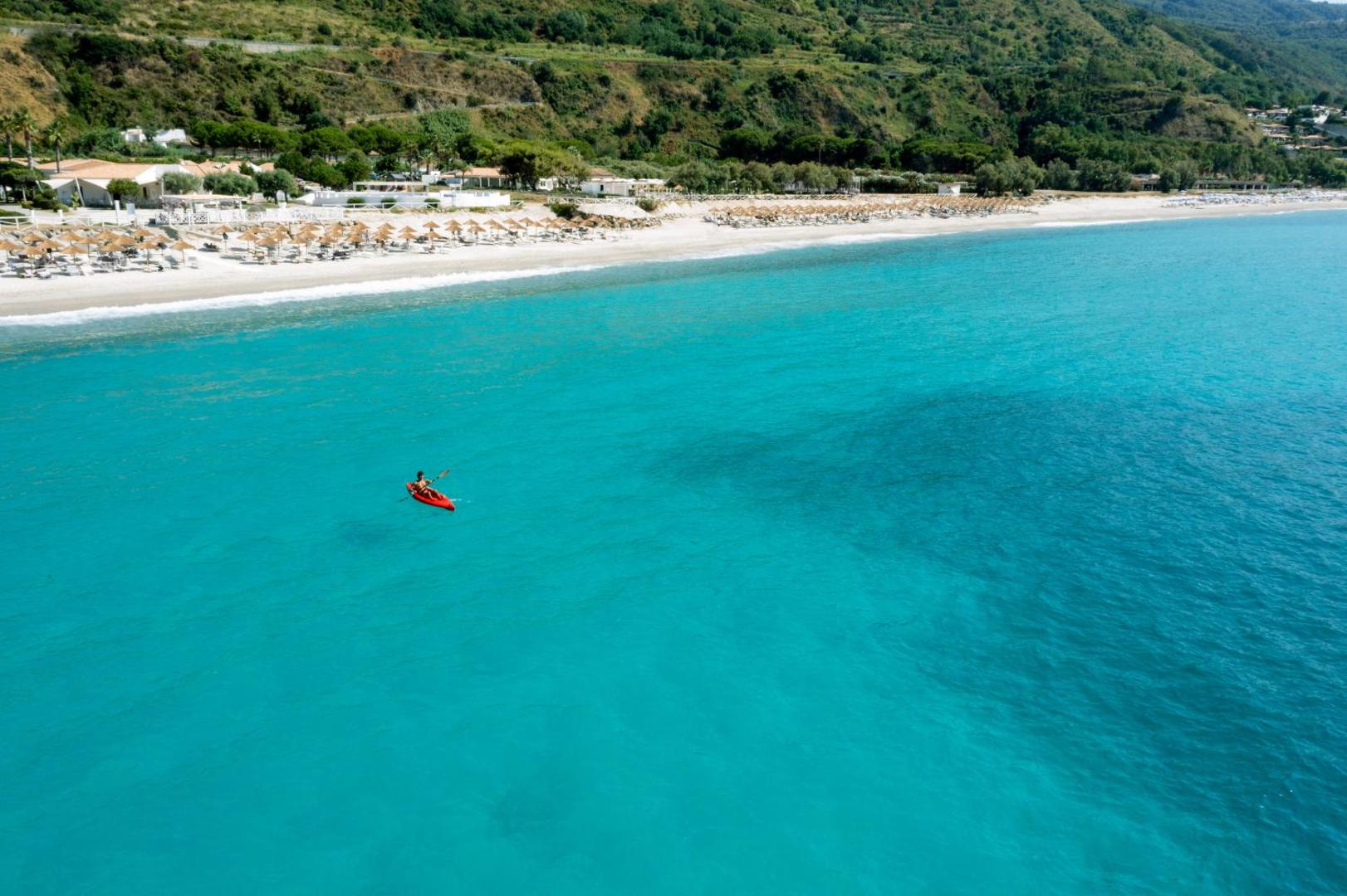 Skats no augšas uz VOI Le Muse Essentia piekrasti ar sarkanu kajaku dzidrā, tirkīzzilā Jonijas jūras ūdenī