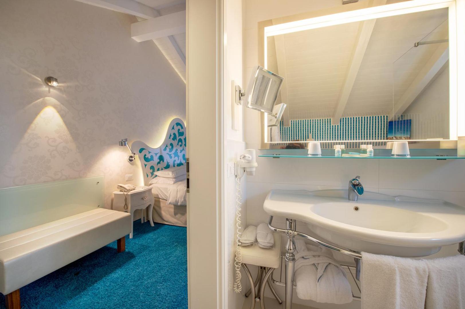 Piccolo Grand Hotel numura interjers ar skatu uz vannasistabu, gaišām mēbelēm un zilu paklāju Kalabrijā