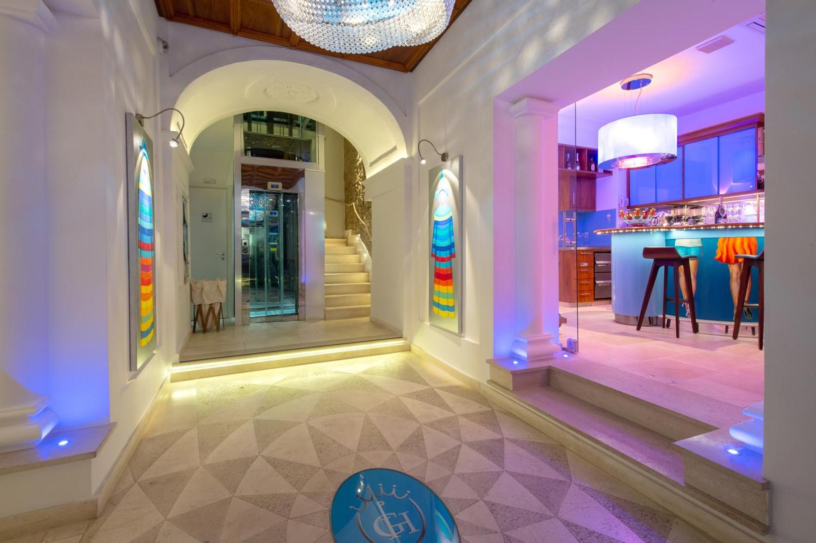 Piccolo Grand Hotel vestibils vakarā ar modernu apgaismojumu, dekoratīvām kolonnām, liftu un ieeju bāra zonā.