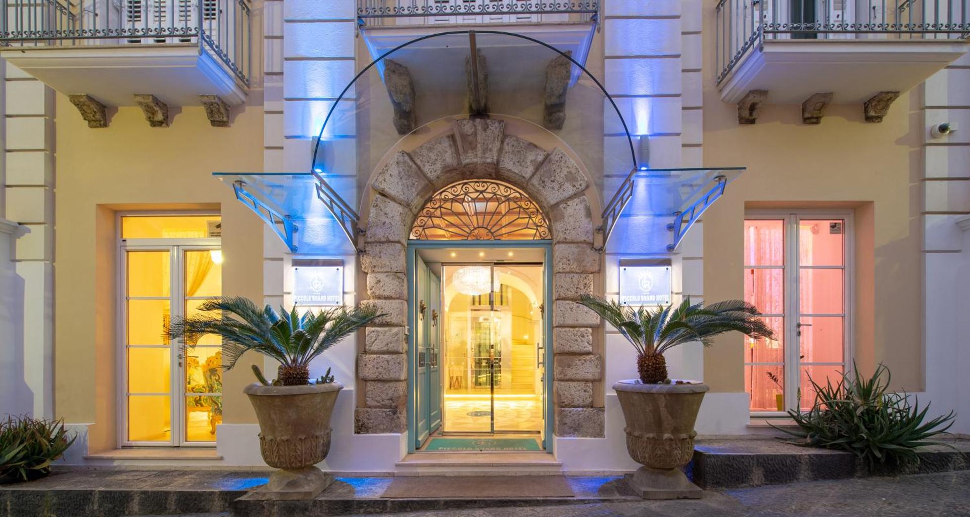 Piccolo Grand Hotel vēsturiskās ēkas ieeja vakarā ar akmens arkām, palmām podos un siltu apgaismojumu