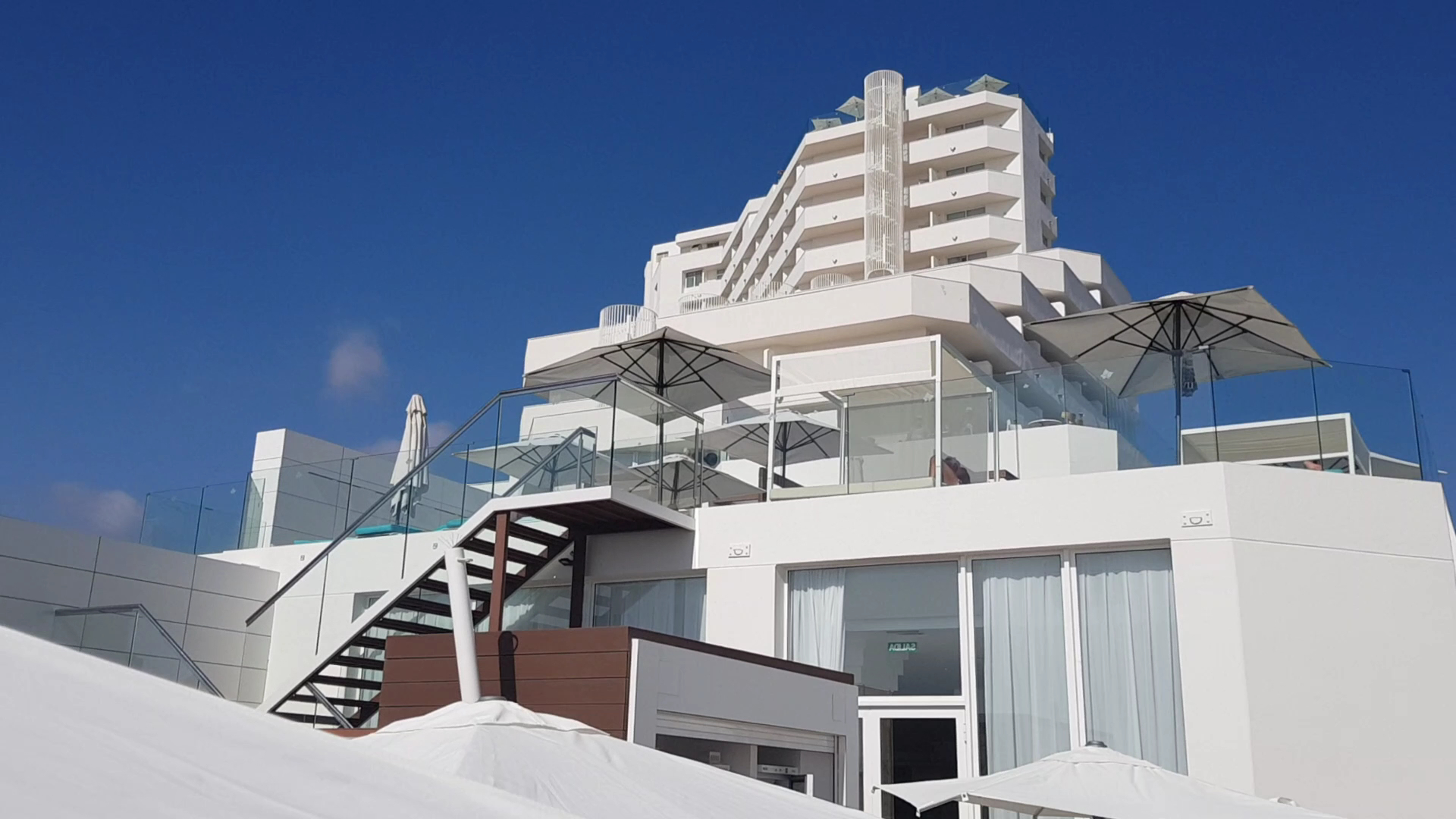 Iberostar Waves Bouganville Playa modernā arhitektūra ar baltām terasēm un saulessargiem pret skaidrām debesīm