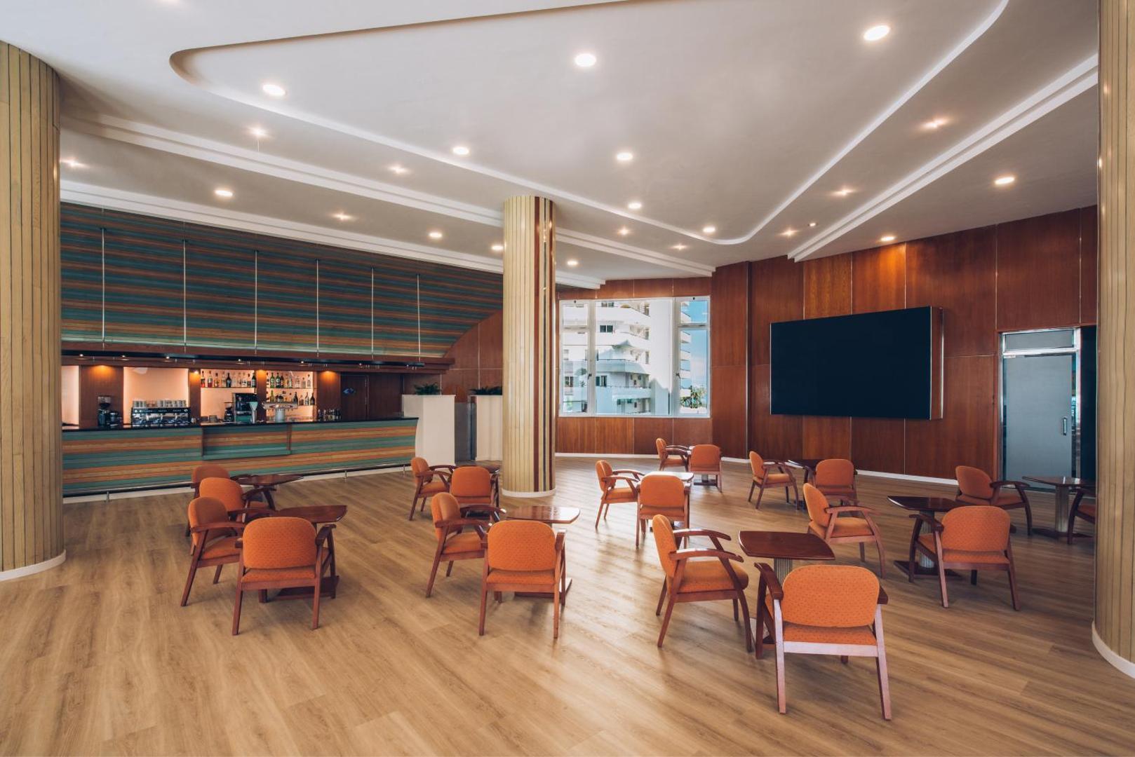 Iberostar Waves Bouganville Playa bārs un atpūtas telpa ar oranžiem krēsliem, koka apdari un lielu ekrānu izklaidei Tenerifē