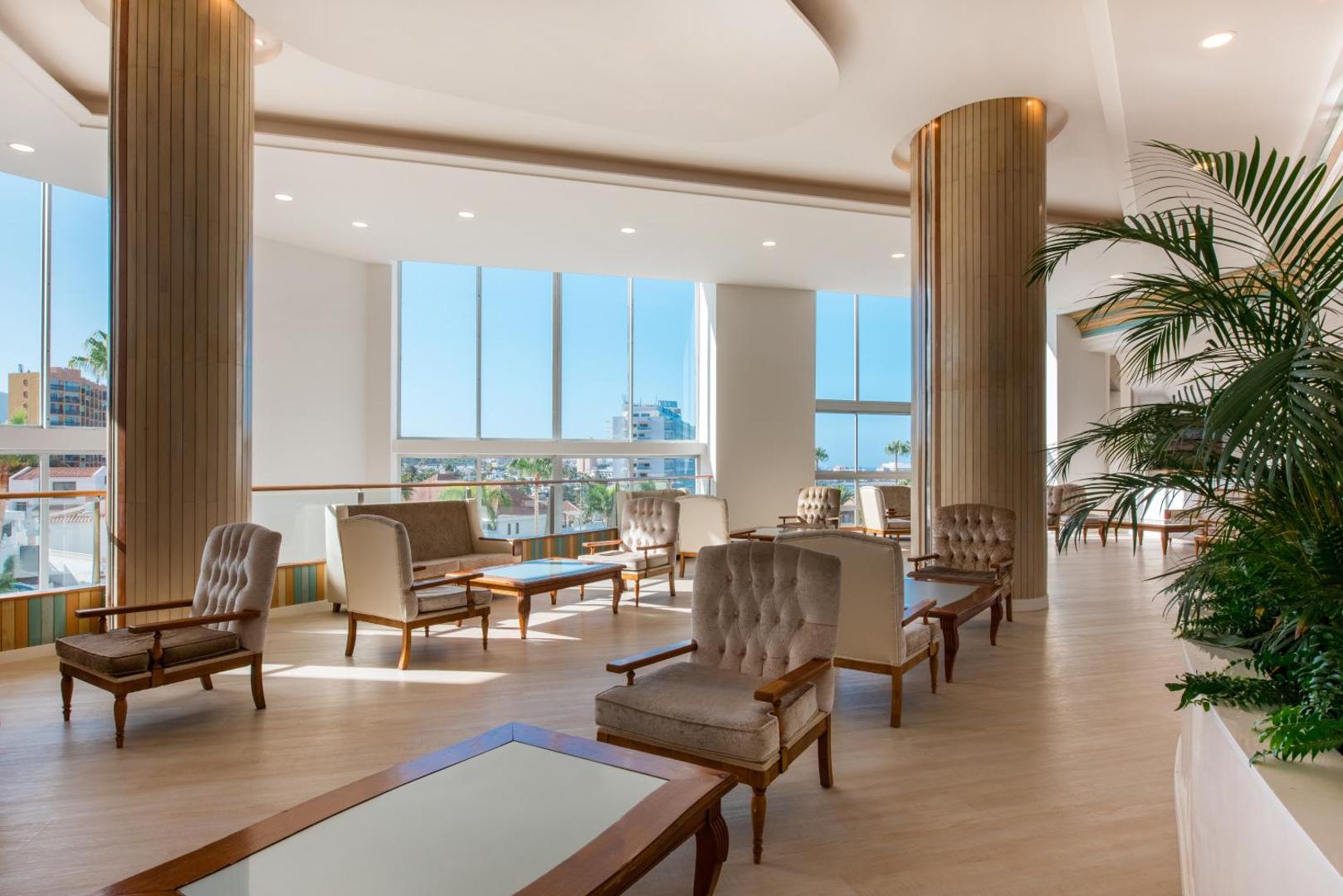 Iberostar Waves Bouganville Playa gaišā lobby atpūtas zona ar mīkstiem krēsliem un skatu uz pilsētu caur lieliem logiem Tenerifē