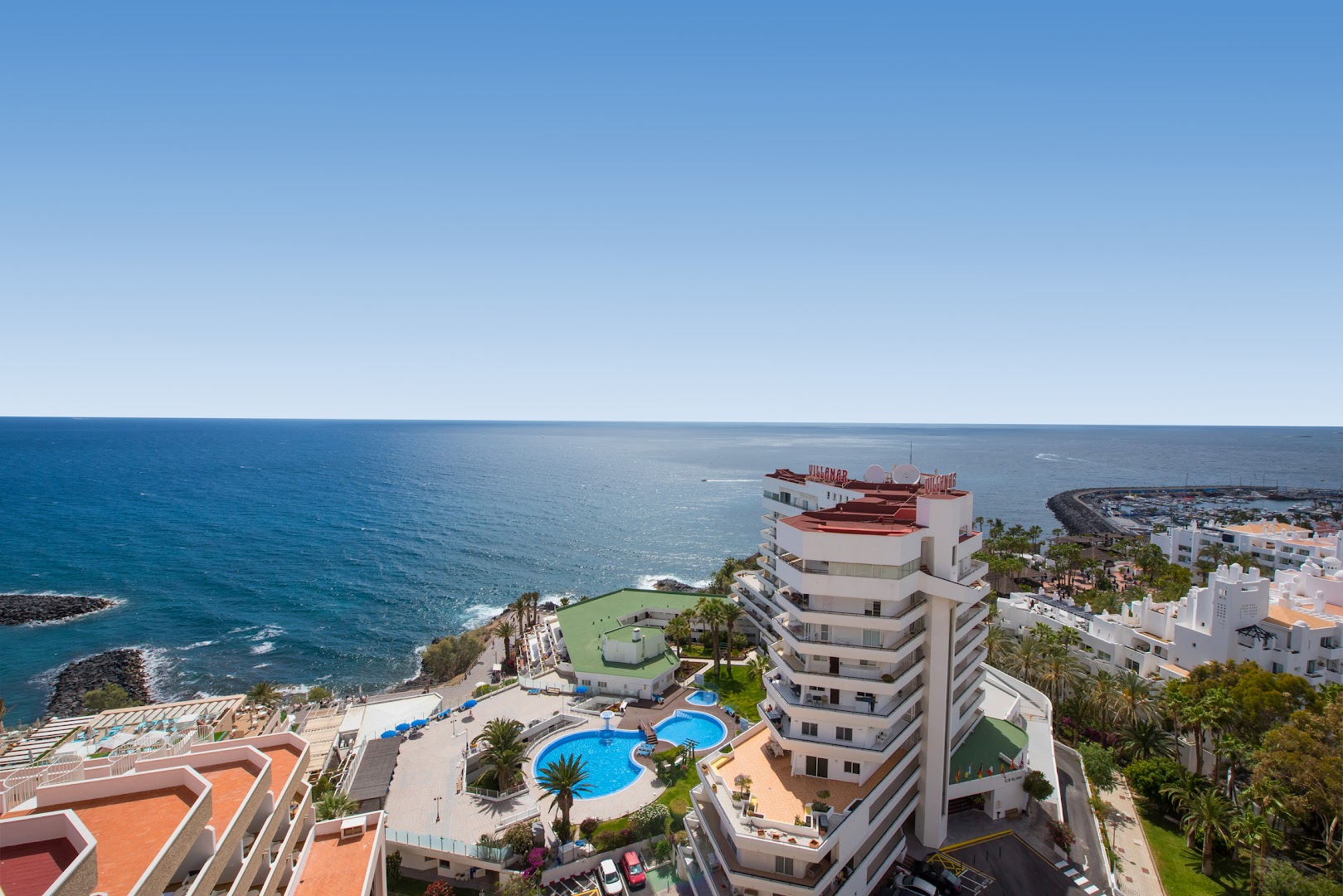 Aerofoto skats uz Iberostar Waves Bouganville Playa kompleksu un zilo okeāna piekrasti ar jahtu piestātni tālumā