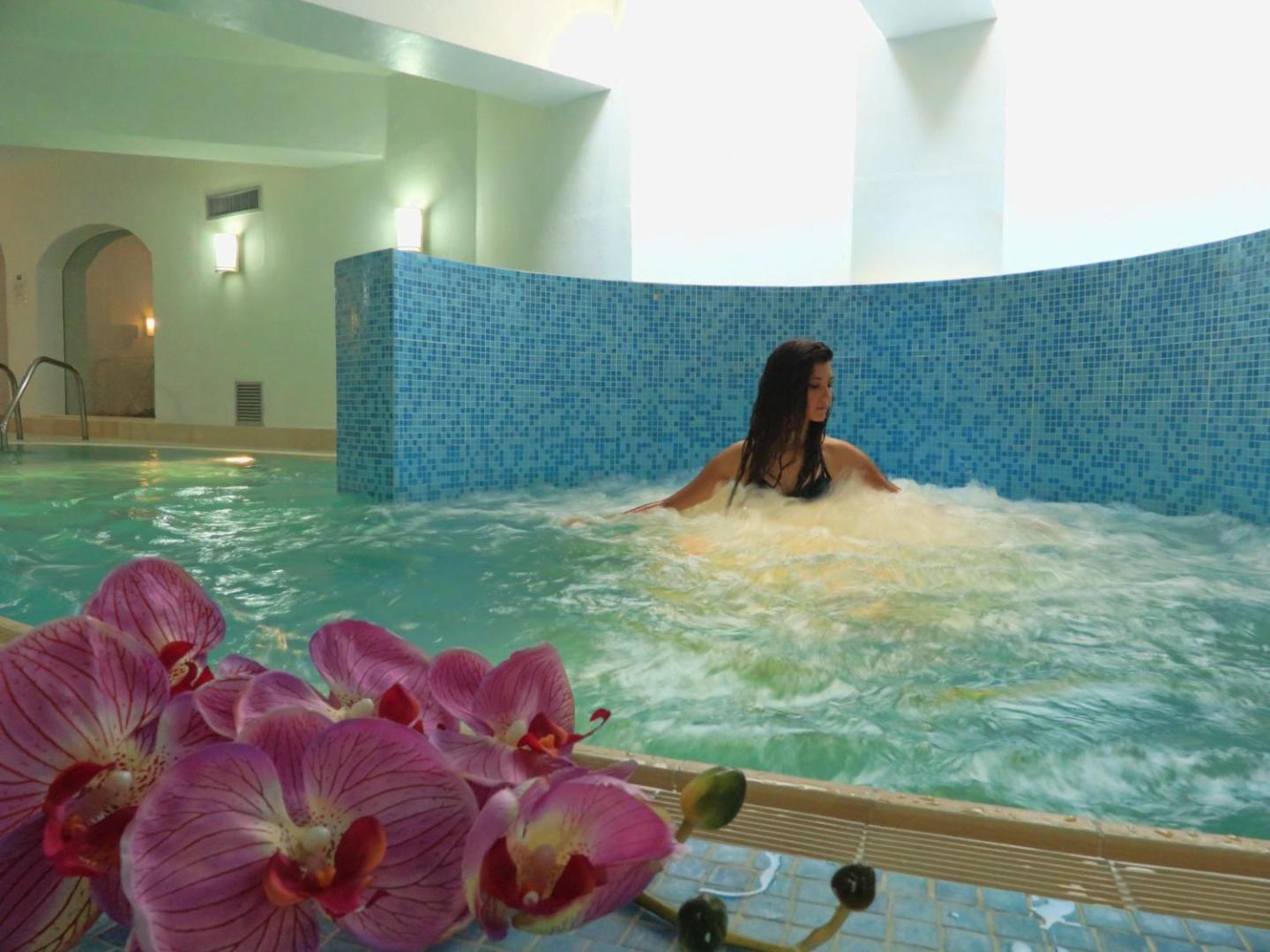Sieviete bauda hidromasāžu Th Le Castella Baia Degli Dei Village spa iekštelpu baseinā ar zilu mozaīkas apdari