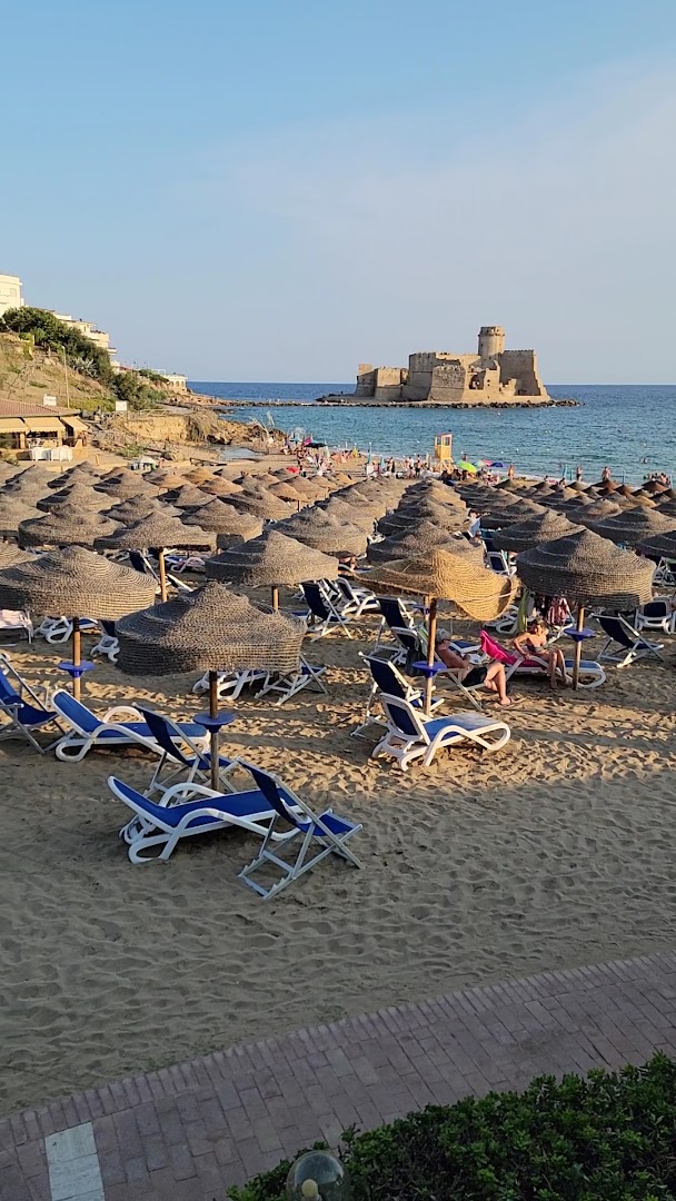 Viesnīcas Th Le Castella Baia Degli Dei Village pludmales zona ar daudziem saulessargiem un zviļņiem, fonā pils jūrā