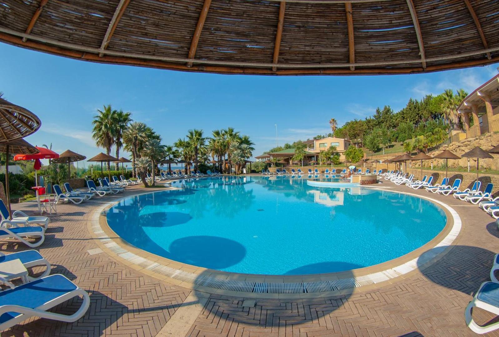 Th Le Castella Baia Degli Dei Village plašais baseins ar saulessargiem un atpūtas krēsliem zem salmu nojumes