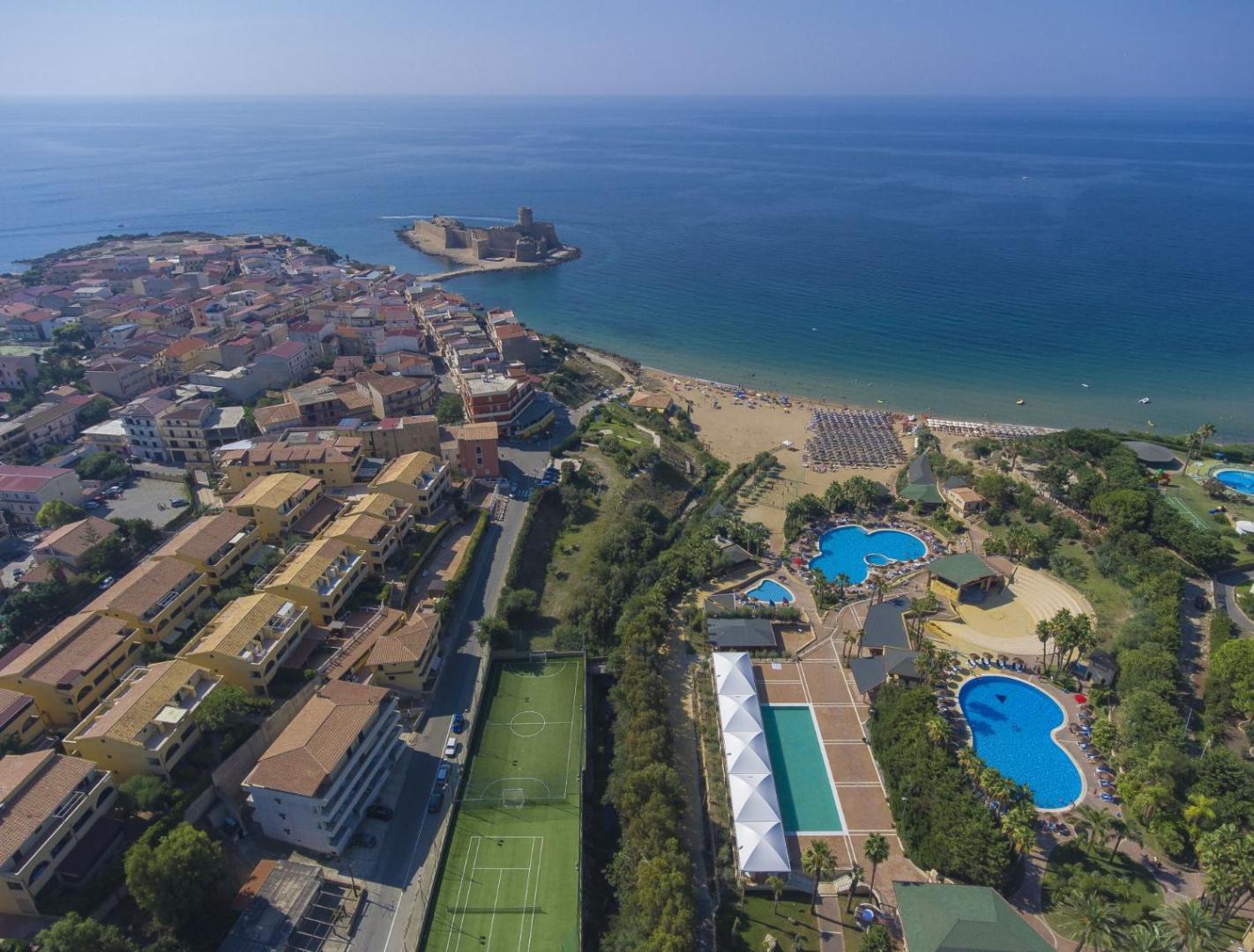 Th Le Castella Baia Degli Dei Village skats no augšas uz kūrorta baseiniem, sporta laukumiem un Le Castella cietoksni fonā