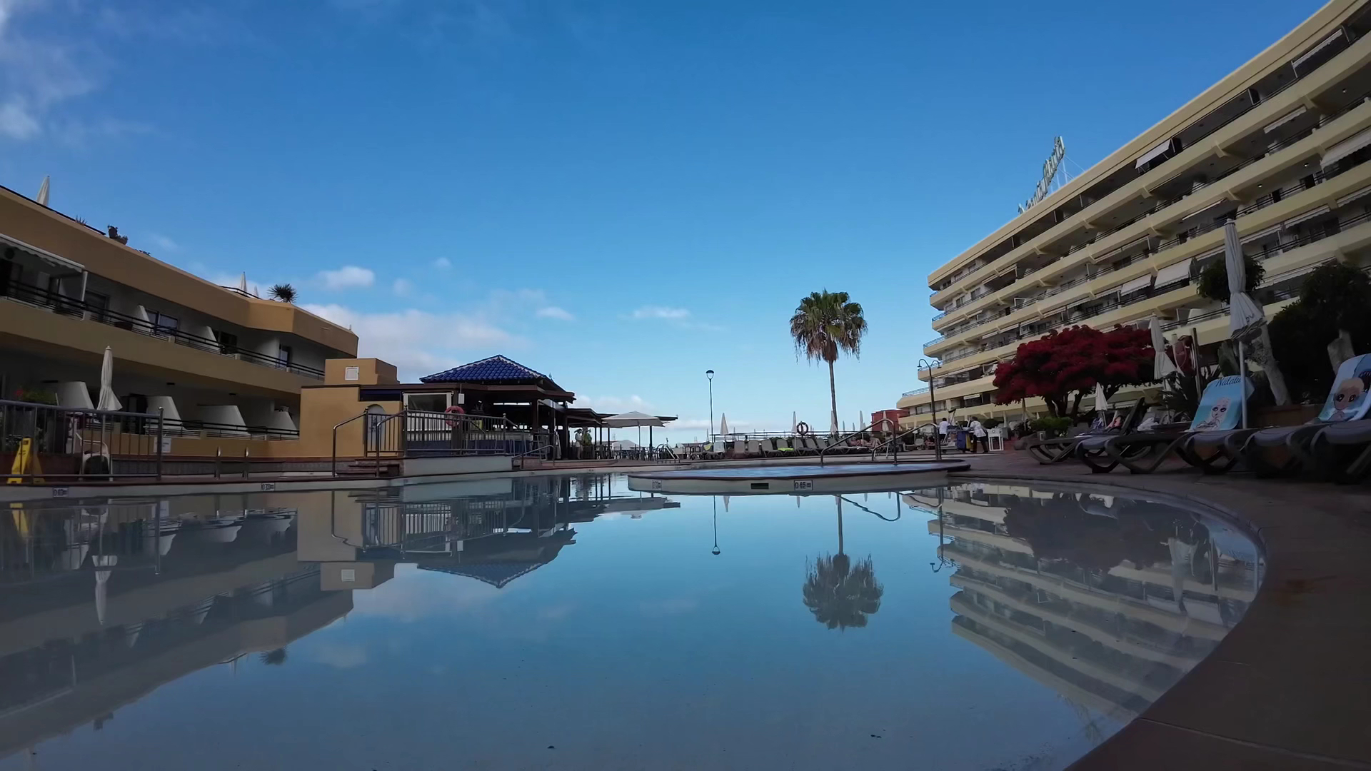 Hovima Santa Maria baseina spoguļattēls mierīgā pēcpusdienā Tenerifē — skats uz viesnīcas fasādi, palmu un zilo debesu atspulgu ūdenī