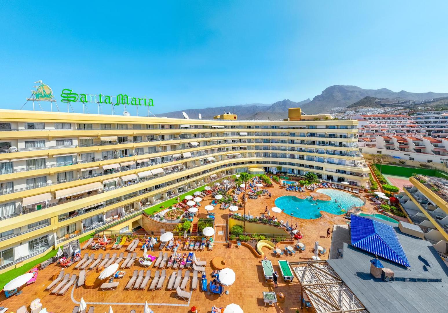 Hovima Santa Maria viesnīcas ēka un galvenais baseins Tenerifē, Kostadeadje — skats uz saulaino terasi ar atpūtas krēsliem un kalniem fonā
