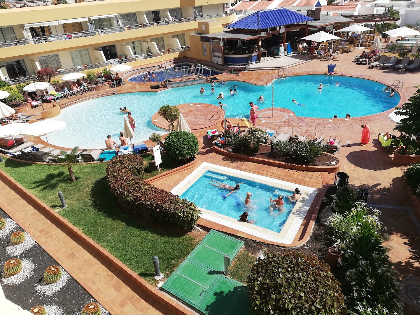 Hovima Santa Maria bērnu un pieaugušo baseini Tenerifē — atpūtnieki bauda sauli, fonā redzams uzkodu bārs un viesnīcas numuru balkoni