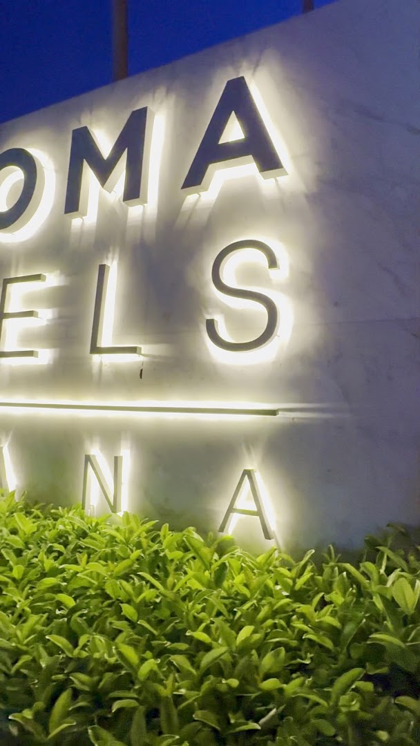 Izgaismots Paloma Hotels izkārtnes uzraksts pie viesnīcas ieejas vakarā, ieskauts zaļos krūmos