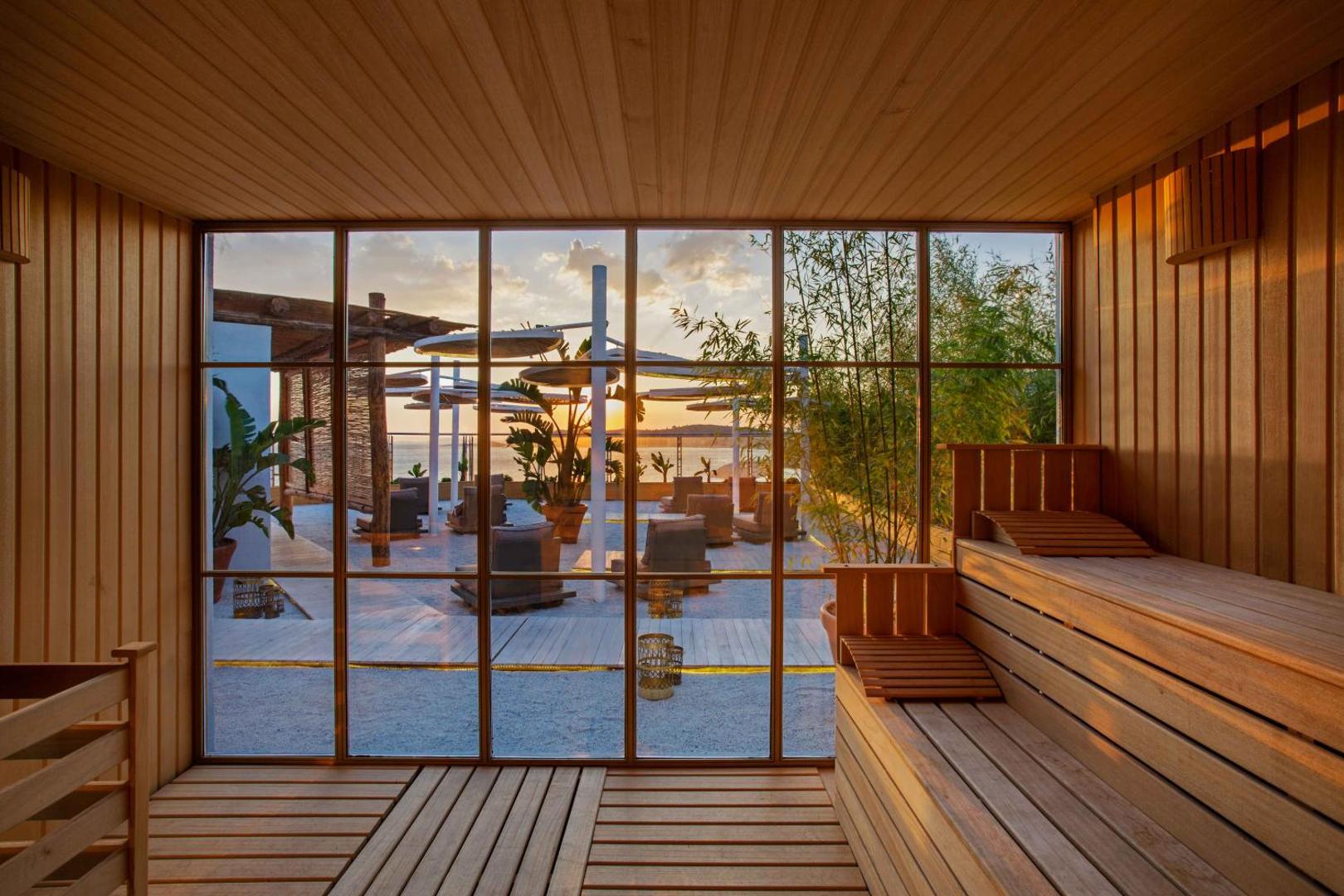 Club Marvy viesnīcas sauna ar panorāmas logu uz āra terasi saulrieta laikā — koka apdare un skats uz jūras piekrasti