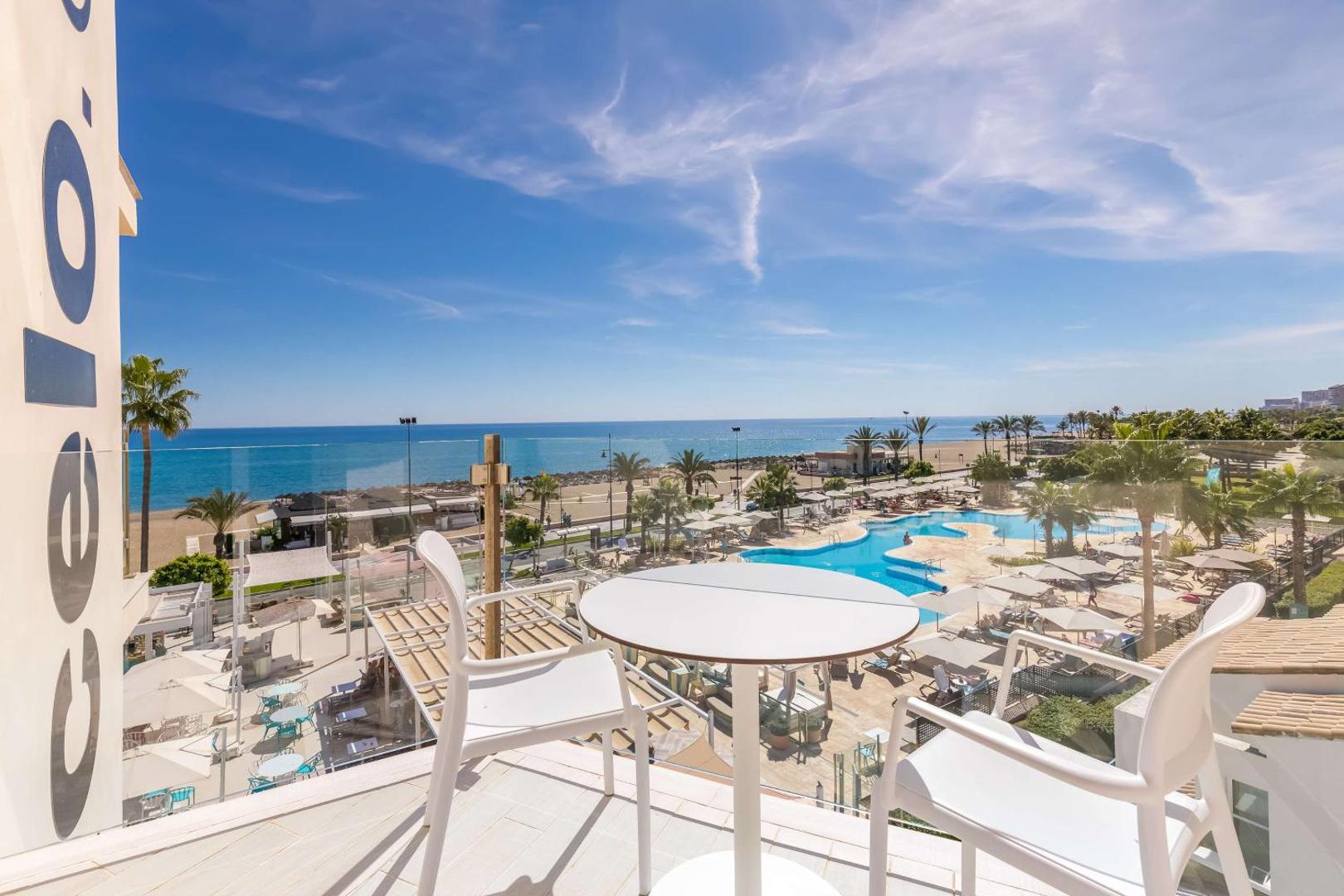 Occidental Torremolinos Playa balkons ar baltu galdiņu un krēsliem, fonā redzams āra baseins, pludmale un zilā Vidusjūra.
