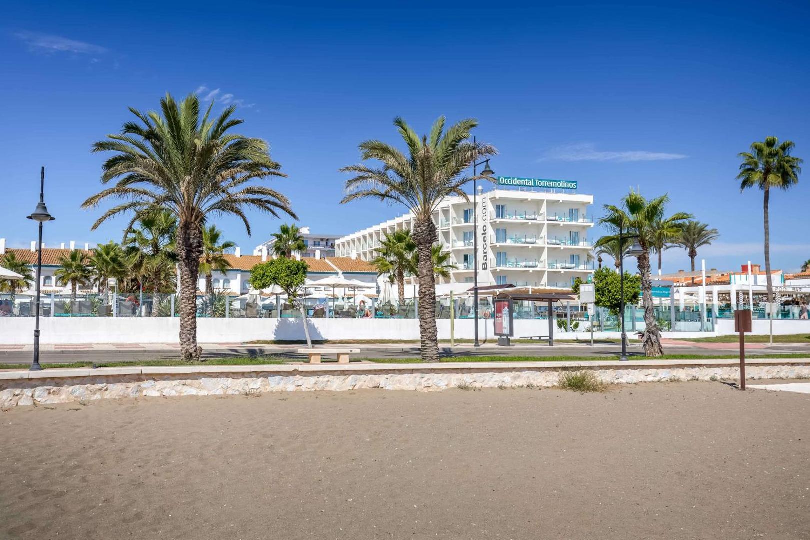 Skats no pludmales smiltīm uz Occidental Torremolinos Playa balto ēku un palmām zem skaidrām debesīm Torremolinosā.