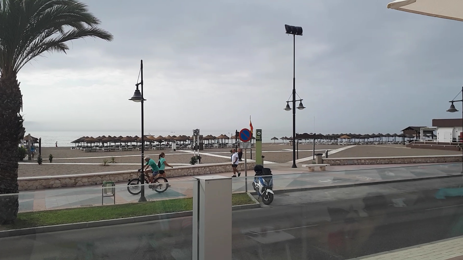 Skats uz Occidental Torremolinos Playa pludmali ar saulessargiem, gājēju promenādi un velosipēdistiem mākoņainā dienā.