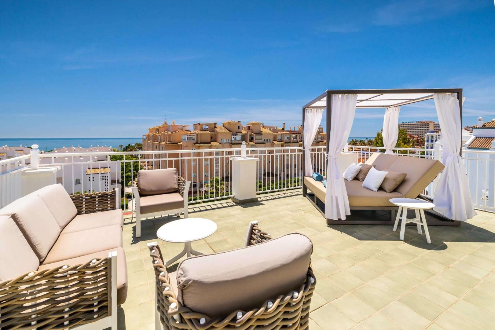 Occidental Torremolinos Playa plaša jumta terase ar atpūtas dīvāniem, galdiņiem un greznu sauļošanās gultu ar skatu uz pilsētu un jūru.