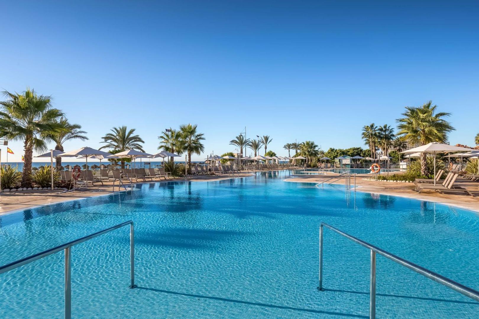 Occidental Torremolinos Playa lielais baseins ar lēzenu ieeju un metāla margām — fonā palmas un rinda ar saulessargiem