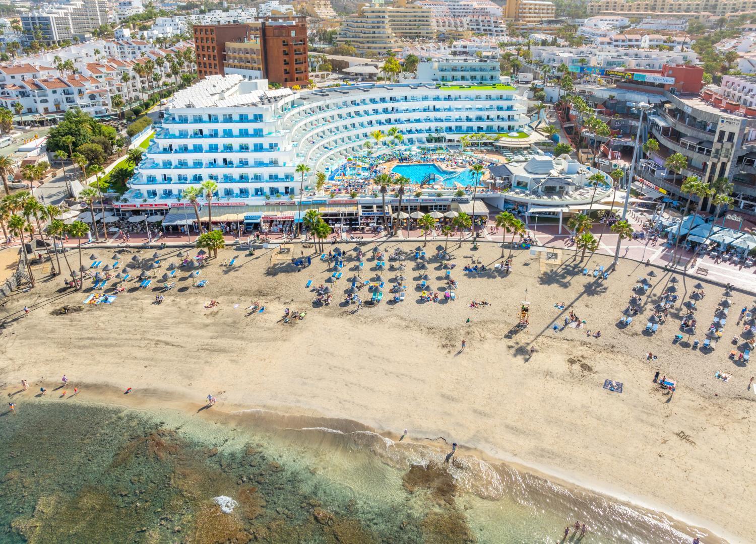 Hovima La Pinta viesnīcas ēka tieši pie smilšu pludmales ar saulessargiem un tirkīzzilu okeāna ūdeni Tenerifes piekrastē