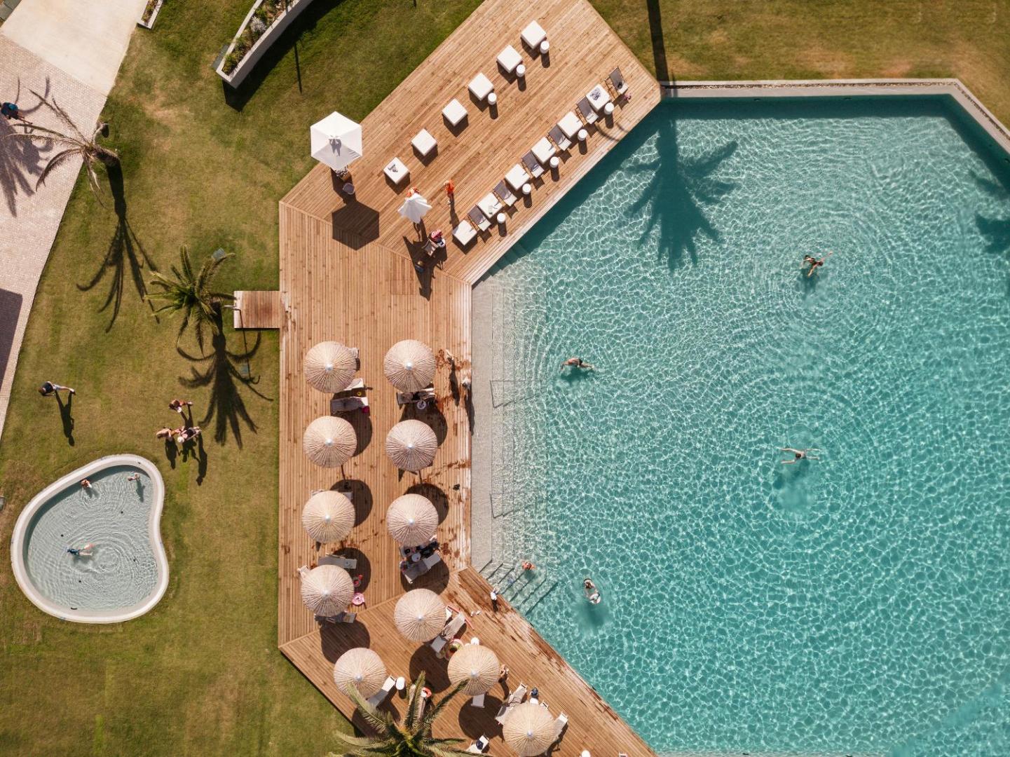 Skats no augšas uz Grecotel Costa Botanica galveno baseinu ar koka klāju, saulessargiem un atpūtas krēsliem Korfu piekrastē.