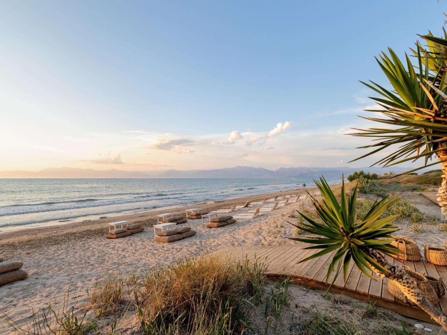 Grecotel Costa Botanica plašā smilšu pludmale saulrietā ar koka laipām un atpūtas vietām — skats uz jūru un kalniem