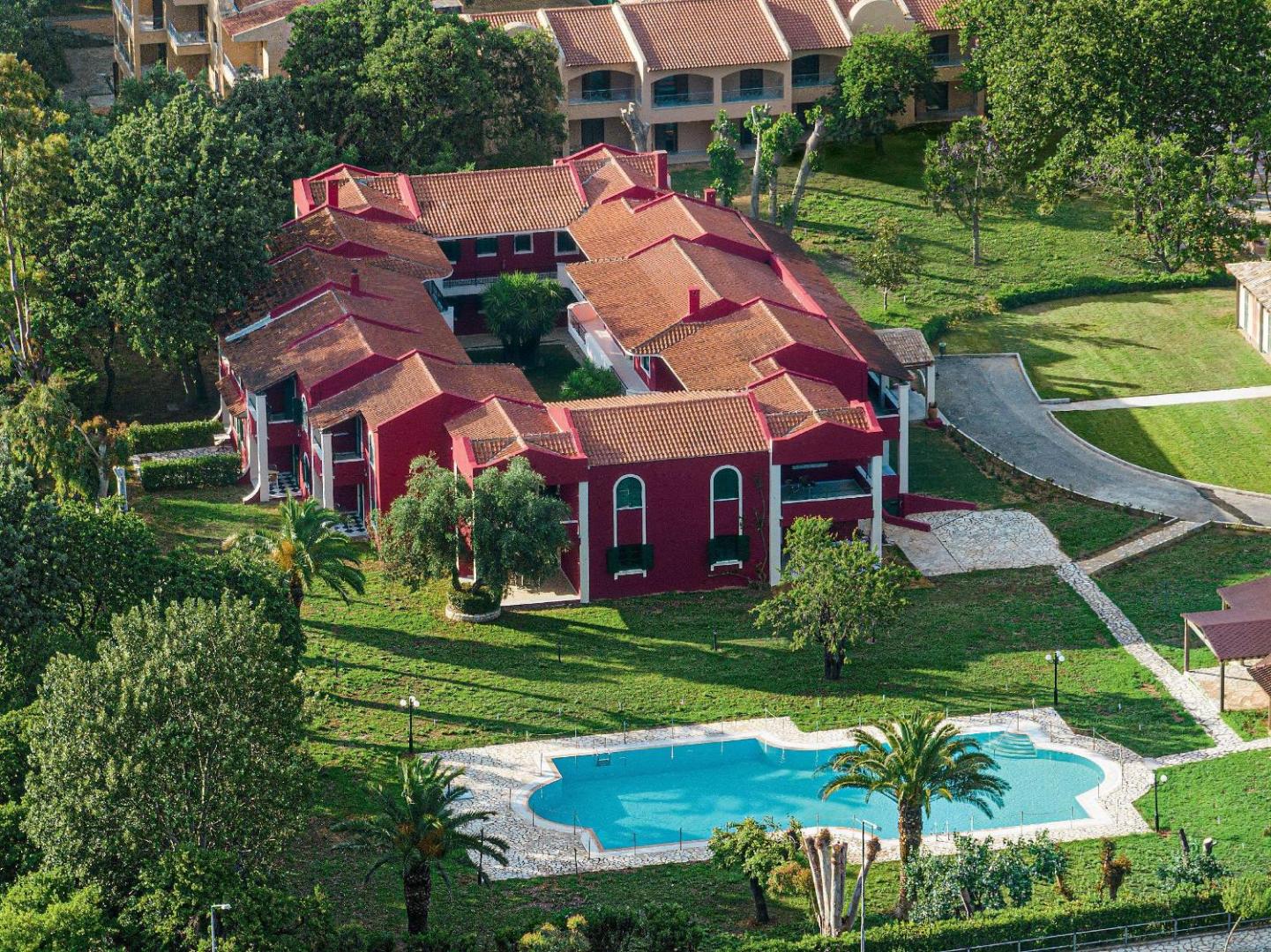 Grecotel Costa Botanica sarkanas krāsas numuru korpuss no putna lidojuma Korfu salā — apkārt zaļš dārzs un neliels āra baseins