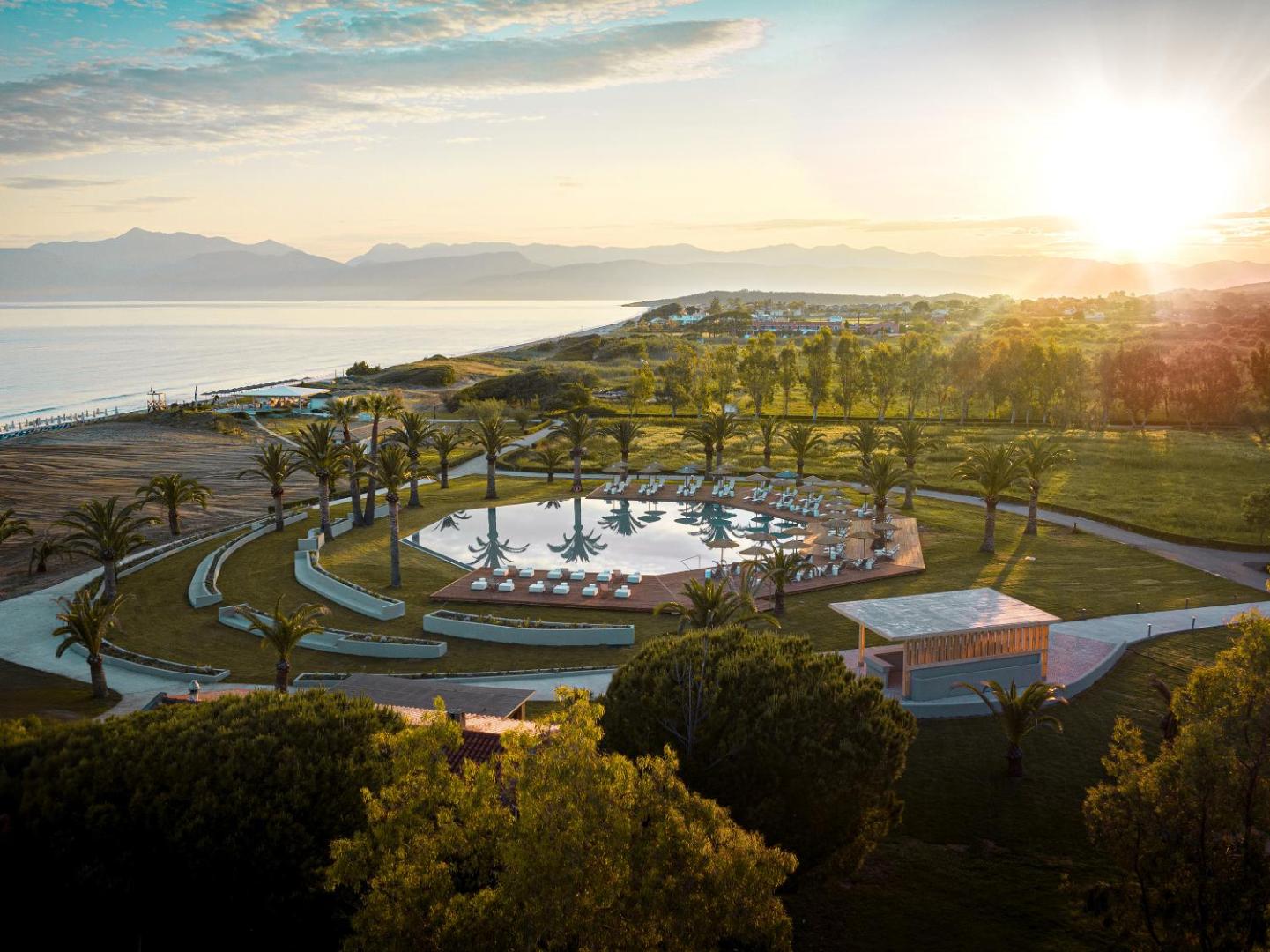 Panorāmas skats uz Grecotel Costa Botanica kūrortu saulrietā ar baseinu, palmām un Jonijas jūru tālumā Korfu salā.