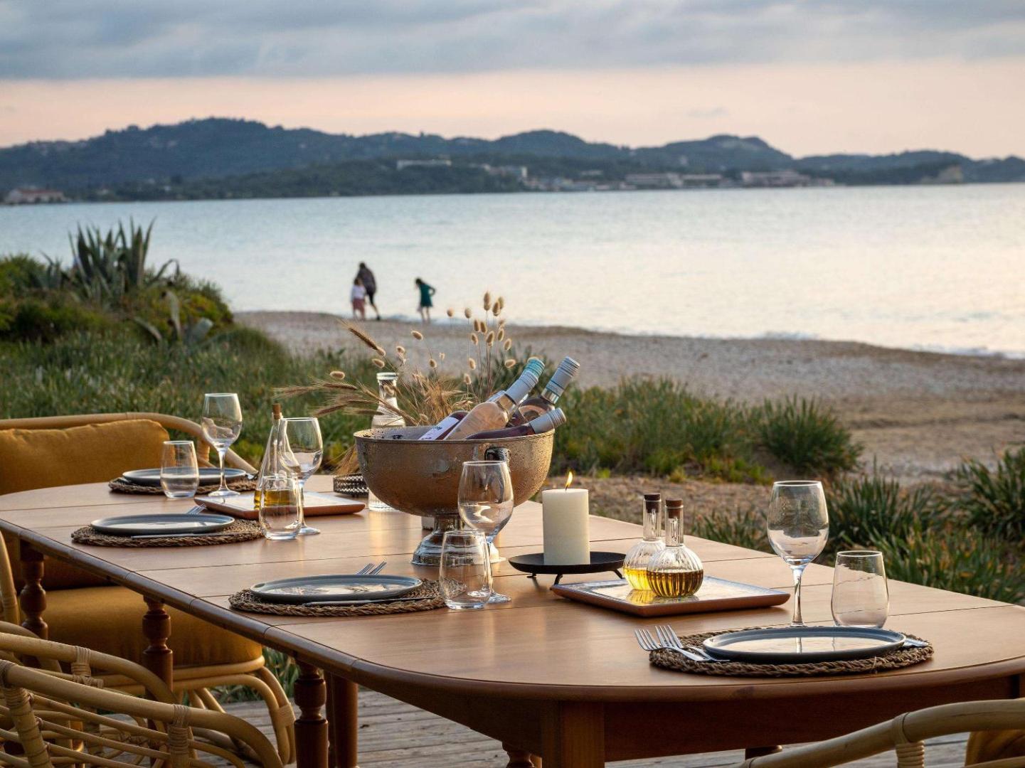 Grecotel Costa Botanica restorāna galds pludmales krastā ar vīnu un svecēm, fonā redzama jūra un kalni saulrietā.