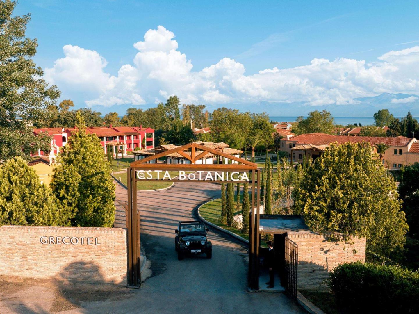 Grecotel Costa Botanica galvenie vārti ar uzrakstu un iebraucamo ceļu — fonā redzami viesnīcas dārzi un kalni tālumā