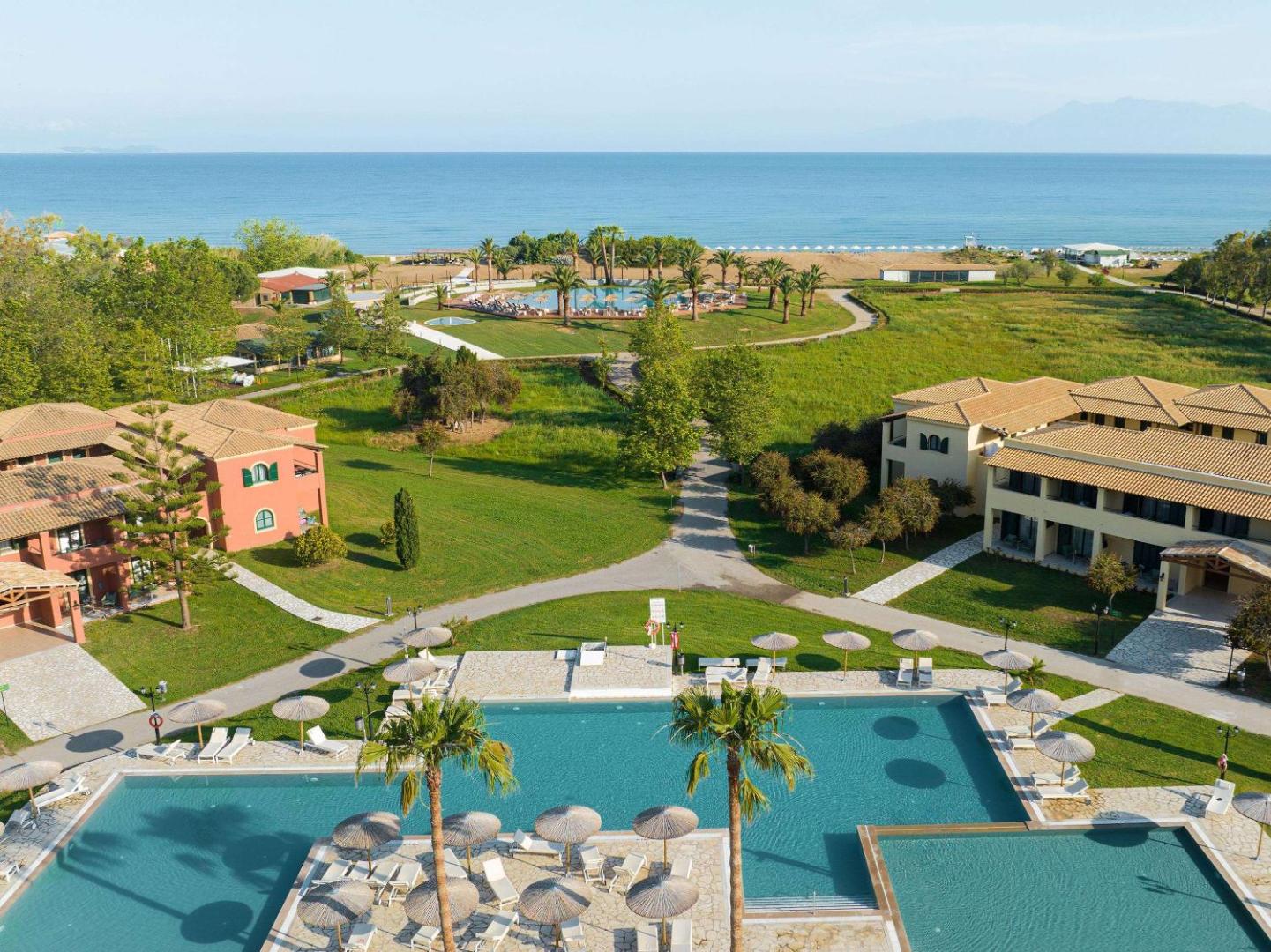 Grecotel Costa Botanica galvenais baseins ar saulessargiem un skatu uz jūru — fonā redzami viesnīcas korpusi un zaļas pļavas