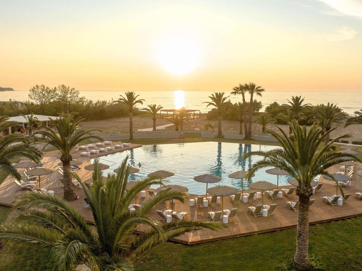 Grecotel Costa Botanica panorāmas skats uz baseinu, palmām un saulrietu pie jūras Korfu salas ziemeļos.