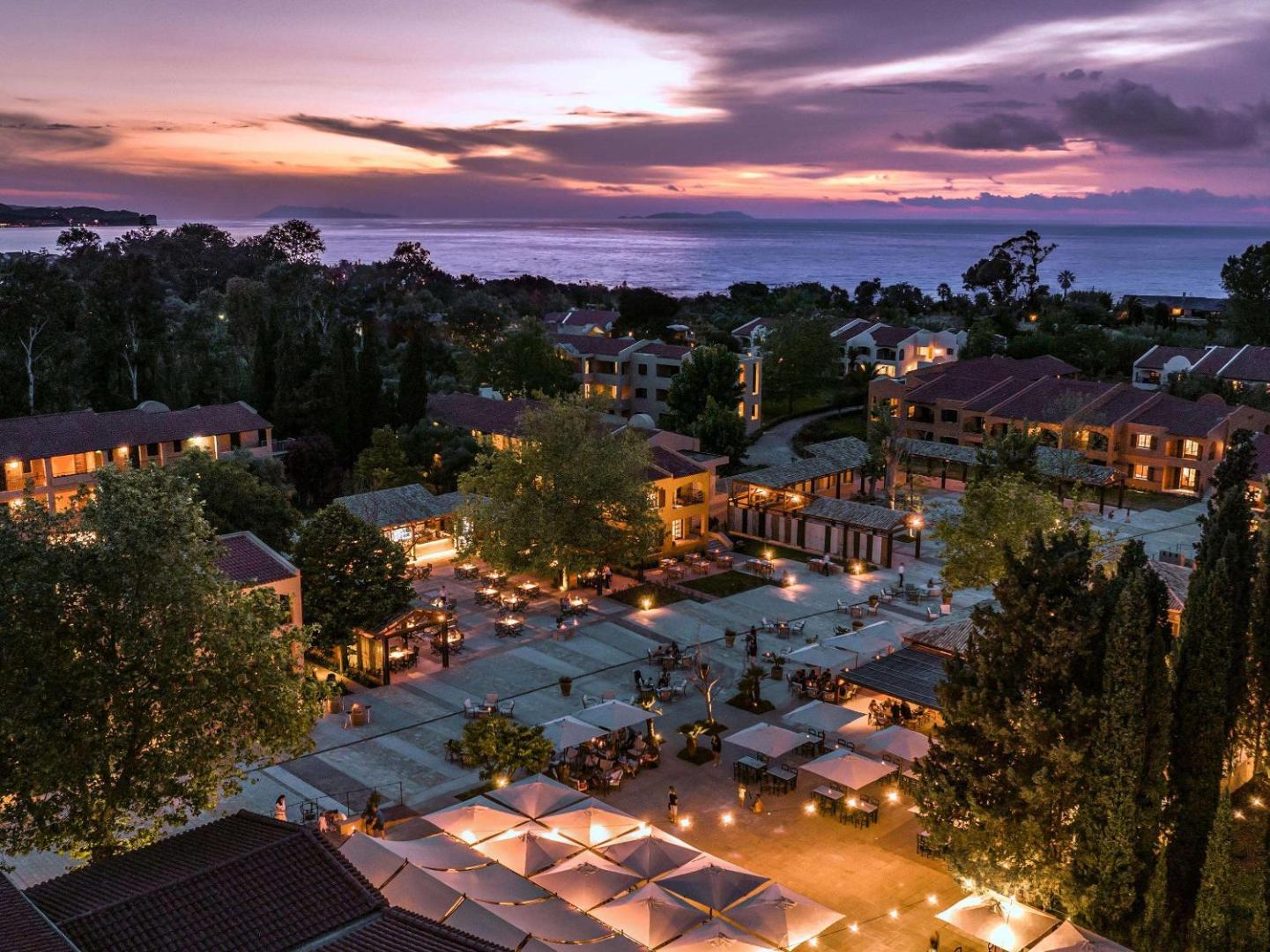 Grecotel Costa Botanica centrālais laukums vakarā ar restorānu galdiņiem un saulrietu pār Jonijas jūru Korfu piekrastē