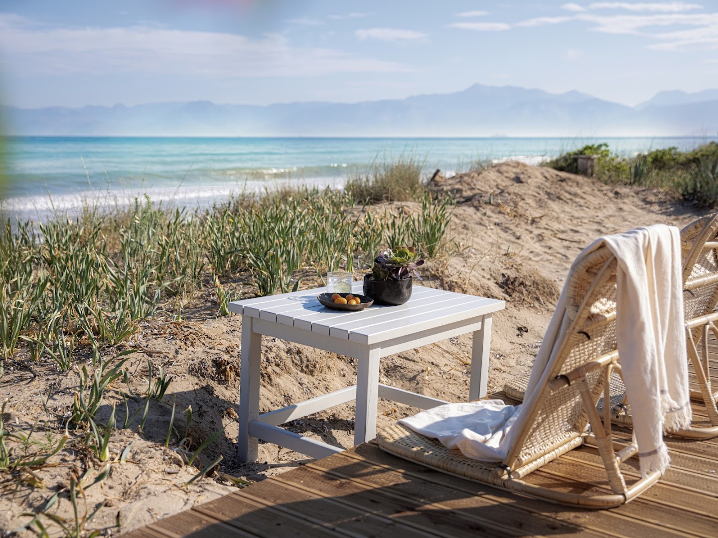 Grecotel Costa Botanica atpūtas stūrītis kāpās ar pītiem krēsliem un galdiņu — skats uz tirkīzzilo jūru un kalniem