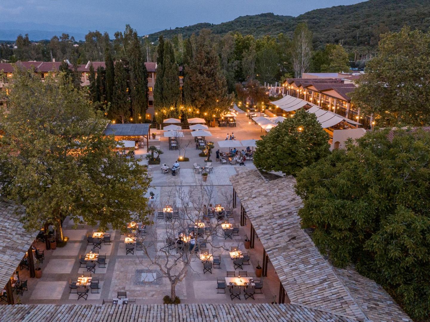 Grecotel Costa Botanica viesnīcas iekšpagalms vakarā ar izgaismotiem restorāna galdiņiem, koku ieskautu terasi un mājīgu apgaismojumu.