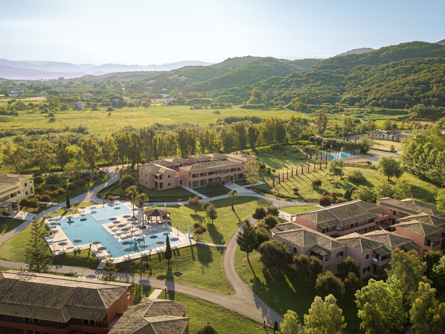 Grecotel Costa Botanica panorāmas skats no putna lidojuma Korfu salā — viesnīcas korpusi, liels āra baseins un zaļojoša kalnu ainava fonā