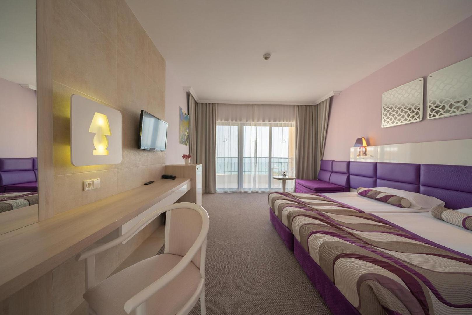 Moderns Grifid Hotel Arabella numurs ar violetiem akcentiem, lielu gultu, dīvānu un stiklotām durvīm uz balkonu