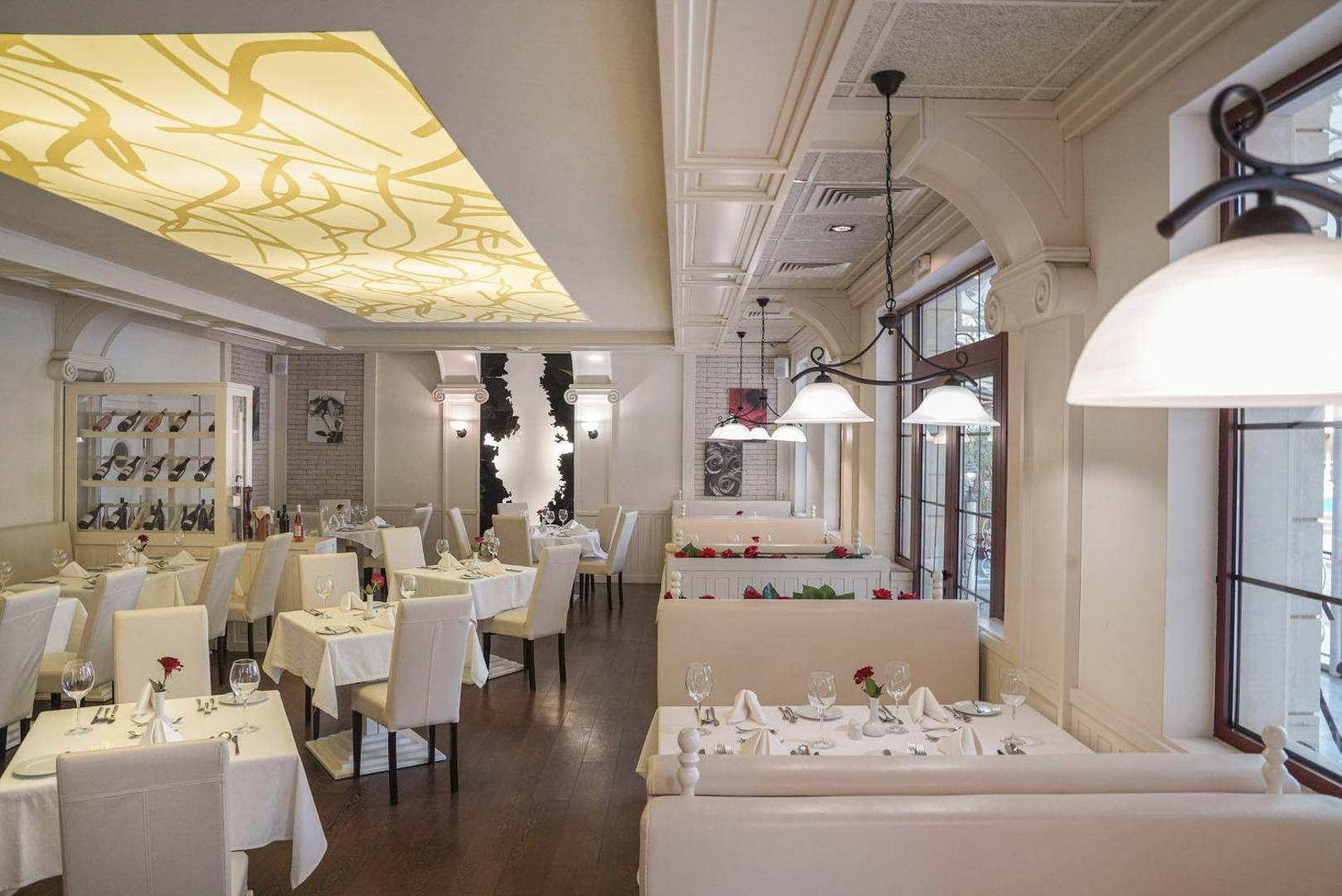 Grifid Hotel Arabella elegants restorāns Zelta Smiltīs ar balti klātiem galdiem, mīkstajiem dīvāniem un dekoratīvu griestu apgaismojumu