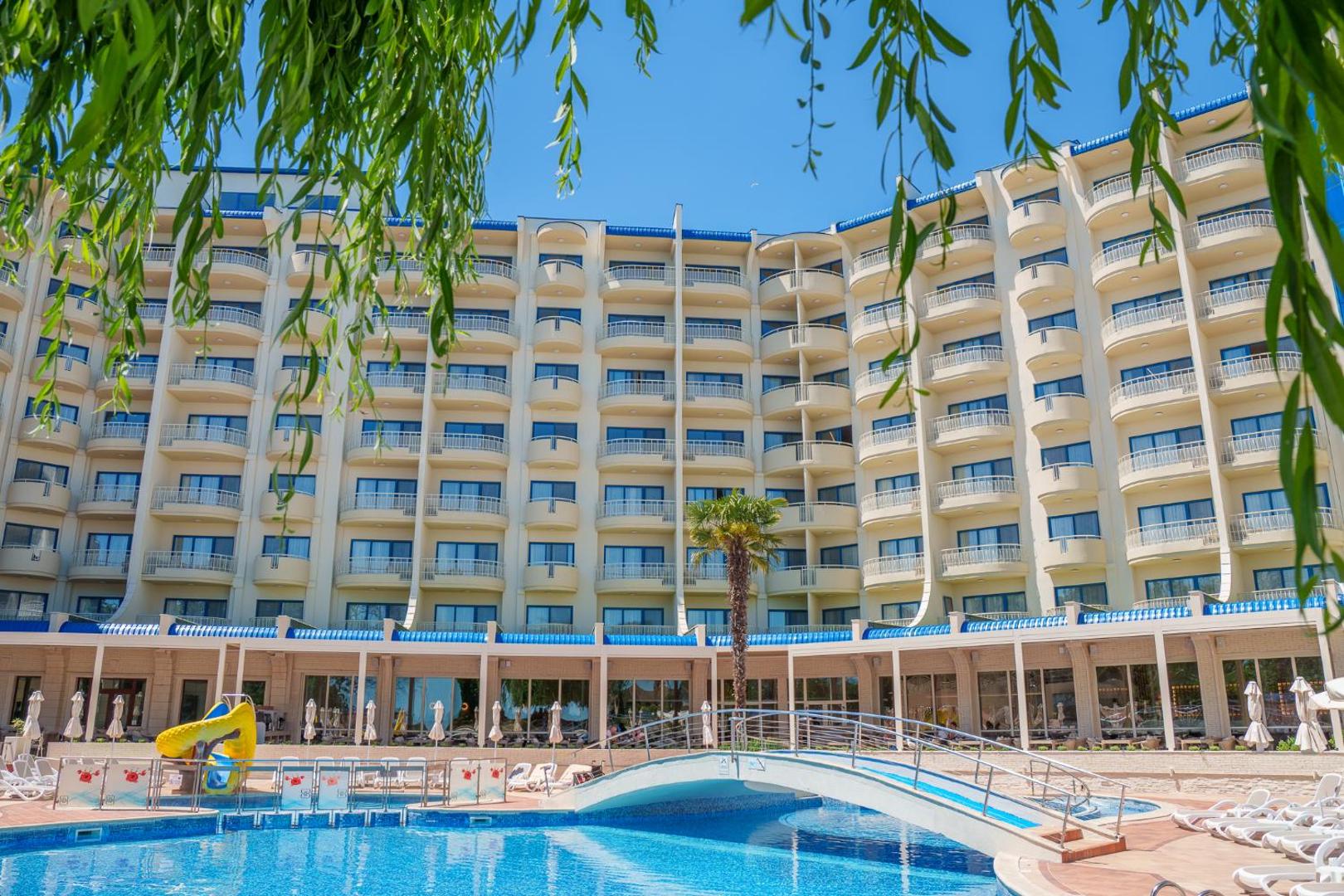 Grifid Hotel Arabella viesnīcas ēka un baseins caur koku lapotni — redzami balkoni, palma un bērnu slidkalniņš