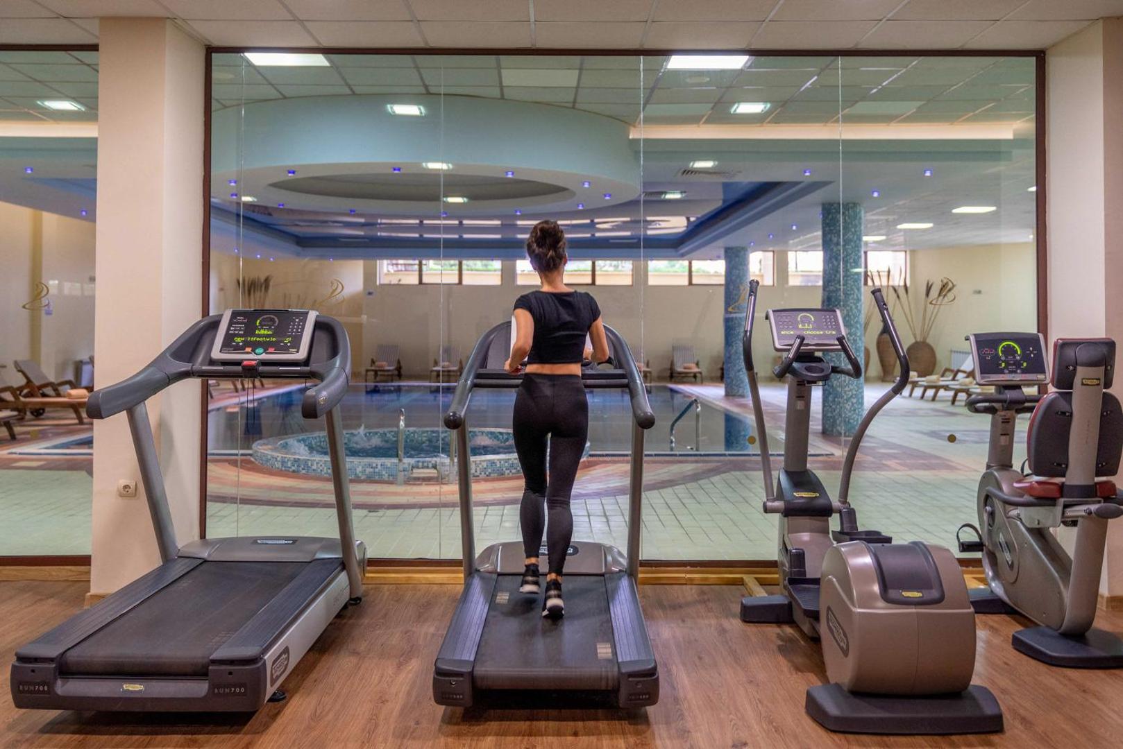 Grifid Club Hotel Bolero fitnesa zāle ar skatu uz iekštelpu baseinu — trenažieri, skrejceliņi un plaši logi sportiskām aktivitātēm