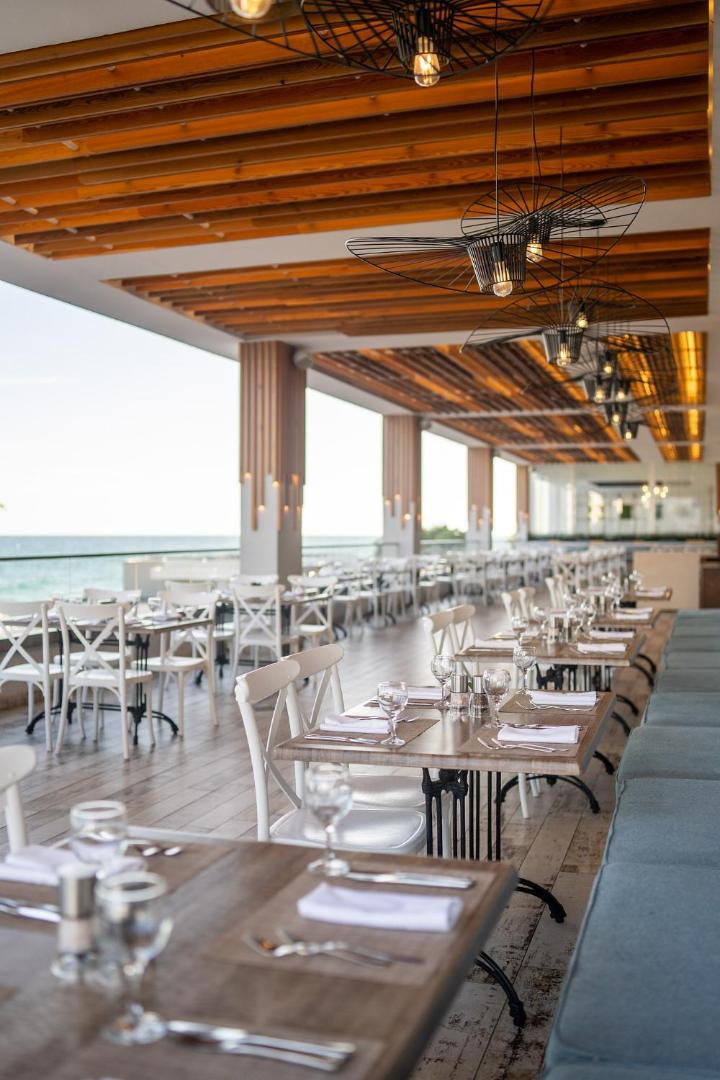Grifid Encanto Beach āra terases restorāns ar baltiem krēsliem, koka galdiem un skatu uz jūru saulrietā
