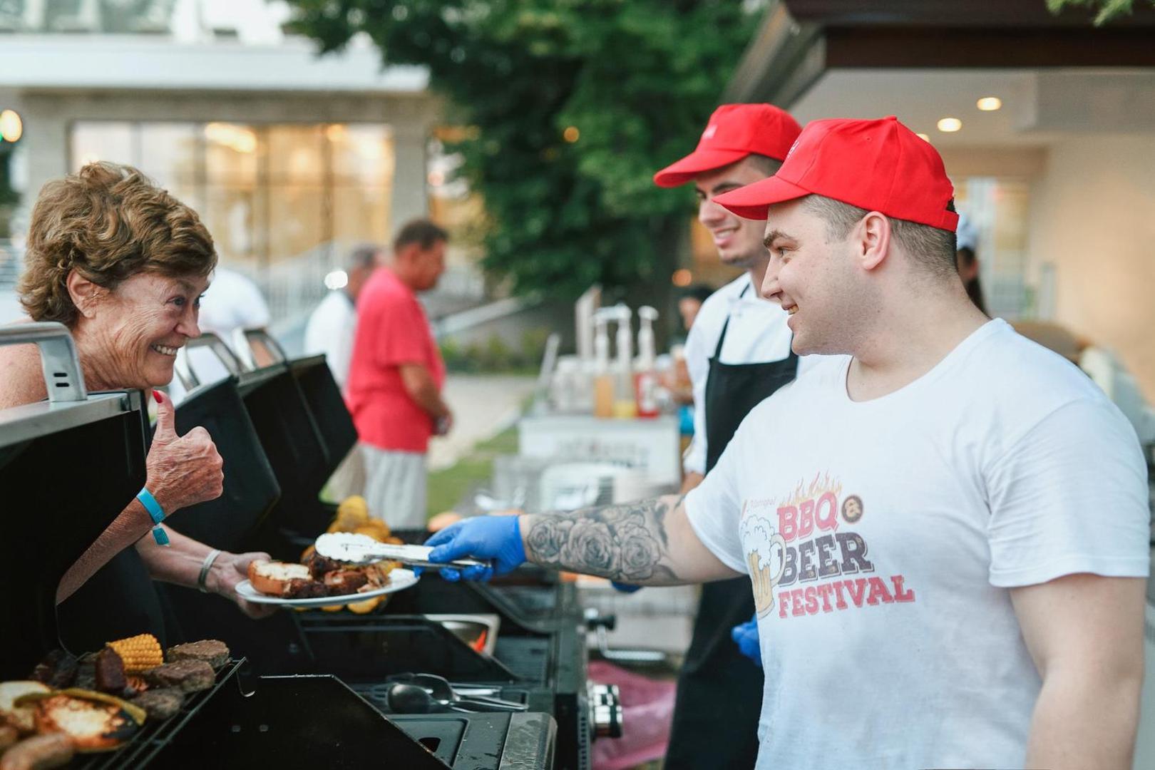 GRIFID Metropol Hotel BBQ un alus festivāls brīvā dabā — pavāri gatavo grilētu gaļu un kukurūzu smaidīgai viesnīcas viešņai