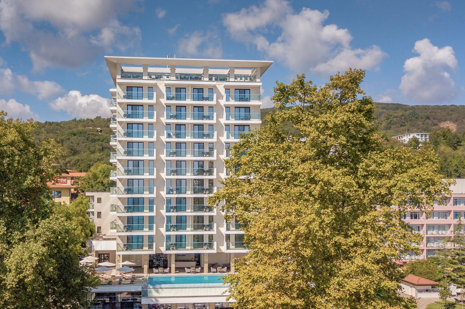 GRIFID Metropol Hotel ēkas skats no priekšpuses ar āra baseinu un saulessargiem — apkārtnē zaļojoši koki un kalni