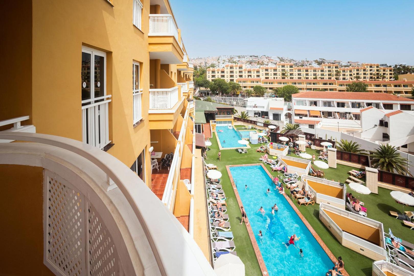 Skats no Villa De Adeje Beach balkona uz diviem āra baseiniem, atpūtas zonu un apkārtējo pilsētas ainavu Tenerifes piekrastē.
