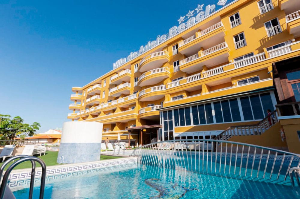 Villa de Adeje Beach viesnīcas ēka ar balkoniem un baseinu priekšplānā — skats no apakšas uz dzelteno fasādi un zilo debesu fonu