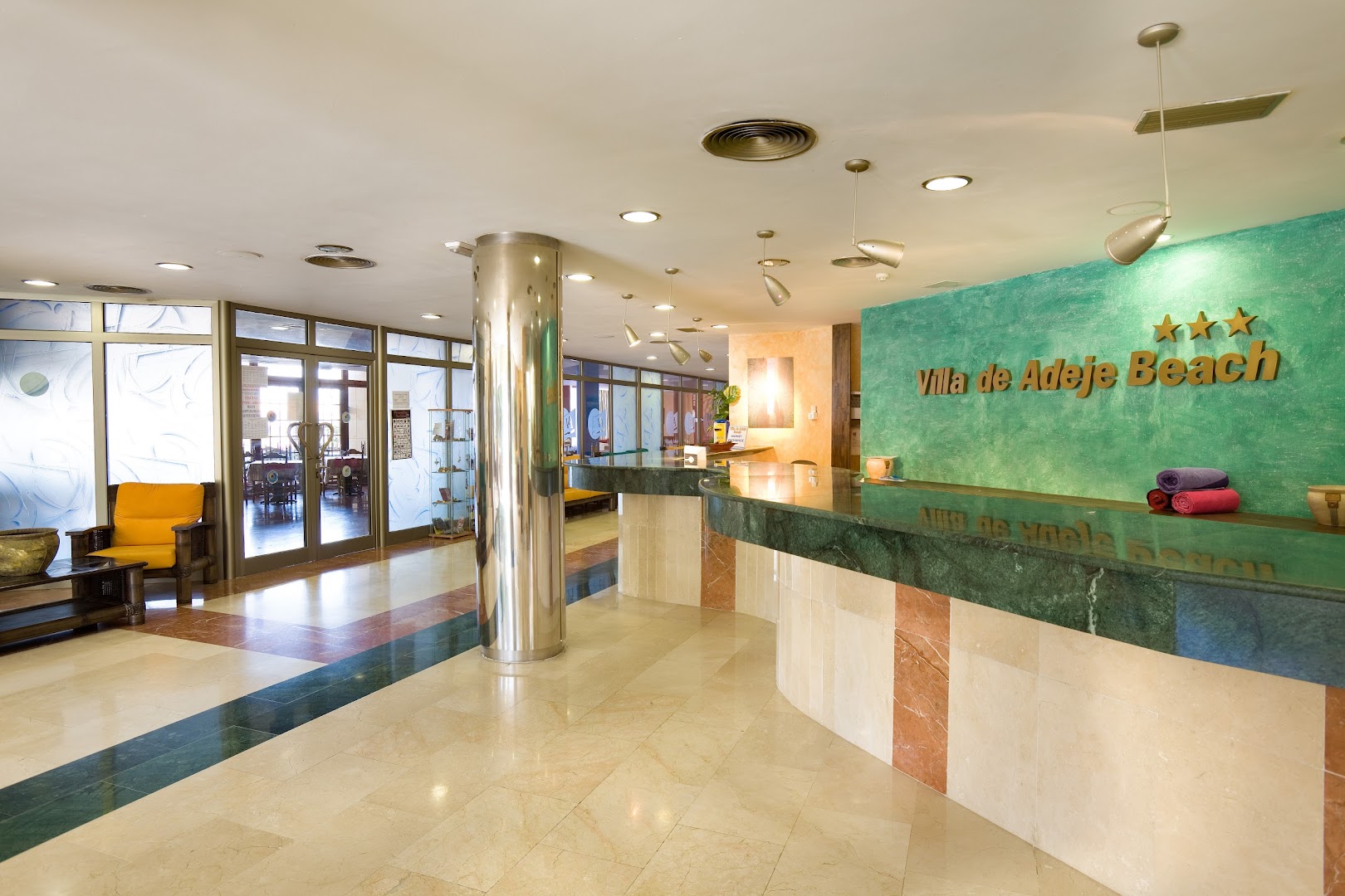 Villa de Adeje Beach viesnīcas recepcijas zona ar marmora grīdu un zaļu akcenta sienu Tenerifē, Spānijā — gaišs vestibils ar atpūtas krēslu