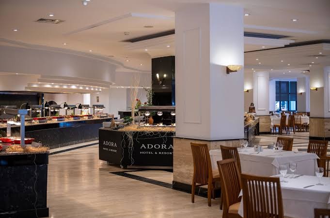Adora Hotel & Resort galvenā restorāna interjers ar klātiem galdiem, koka krēsliem un plašu bufetes līniju fonā