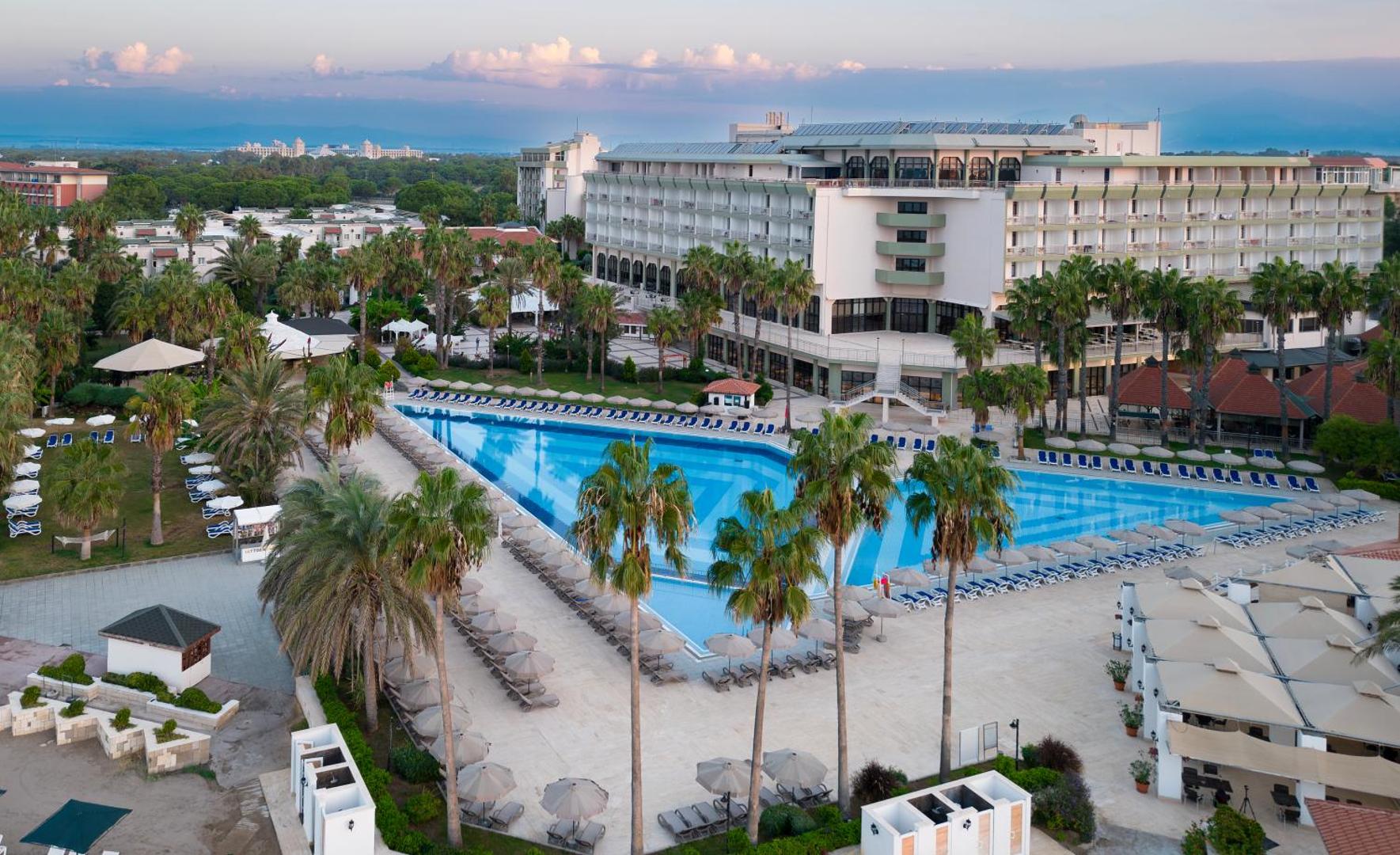 Adora Hotel & Resort centrālais baseins ar ziliem rakstiem vakarā — apkārtnē palmas, atpūtas krēsli un viesnīcas ēka saulrietā